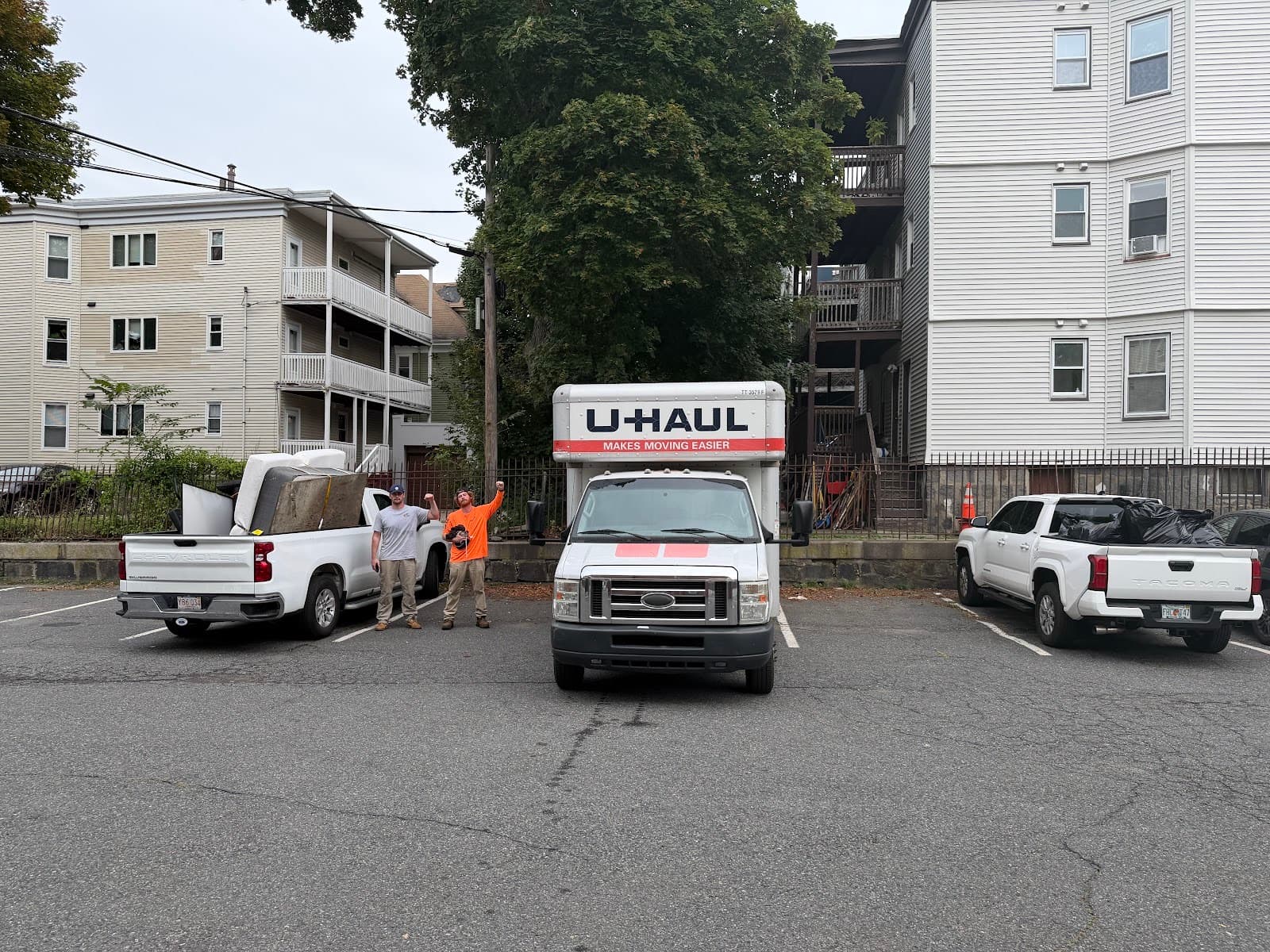 Two Harbor Haul crew members waving beside two pickup trucks and a U-Haul box truck in a parking lot
